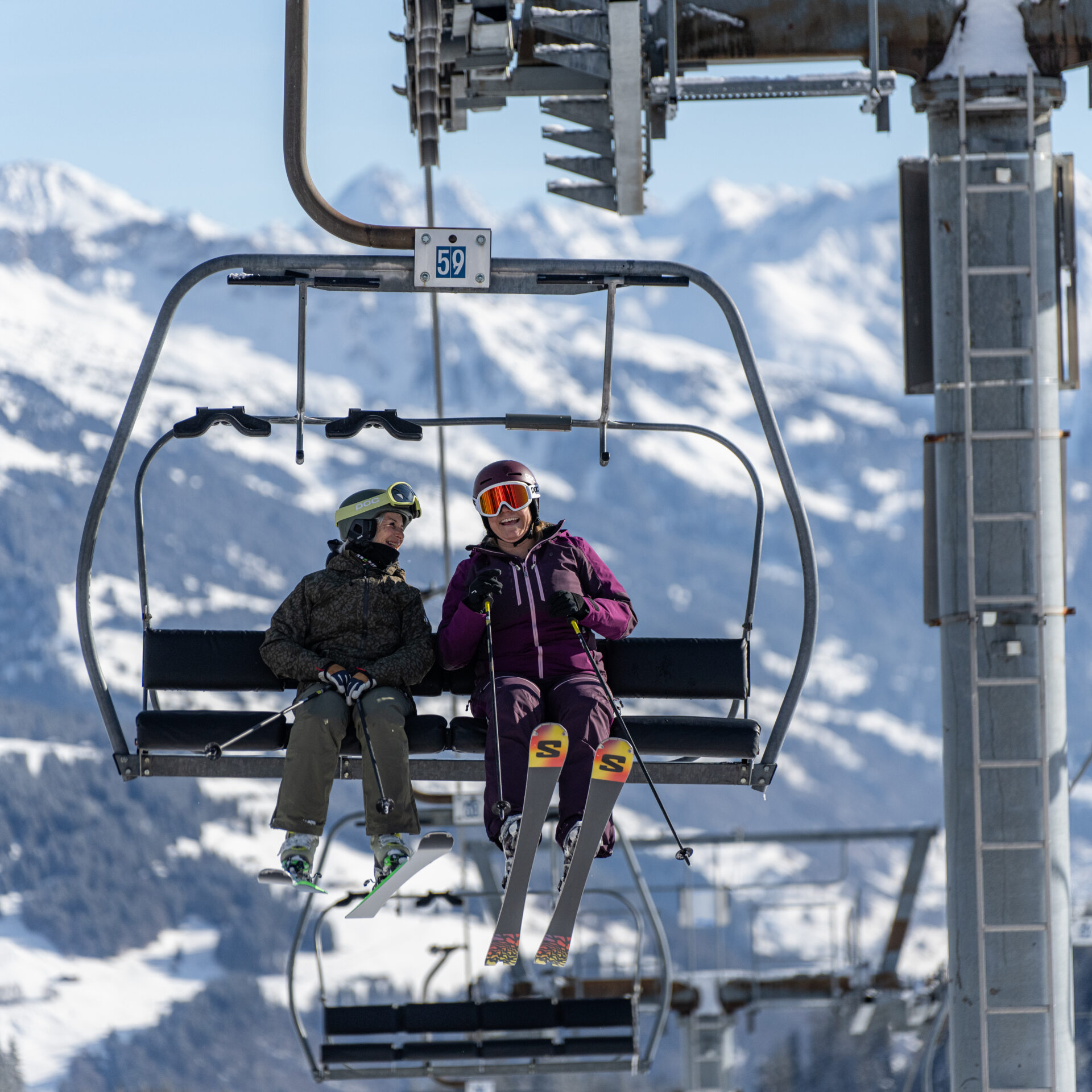 Zwei Frauen sitzen auf dem Sessellift und lachen über etwas Gesagtes. Dahinter verschneite Berglandschaft